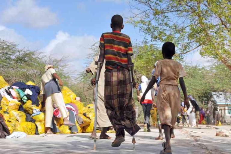 A child with crutches walks beside another child on a sunny street, with people sorting through yellow bags in the background.