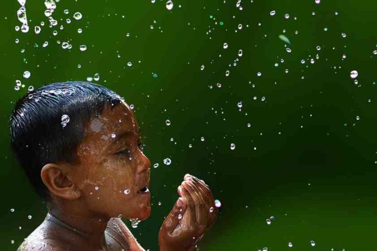 A child with wet hair raises a hand to their mouth while standing among falling water droplets against a green background.