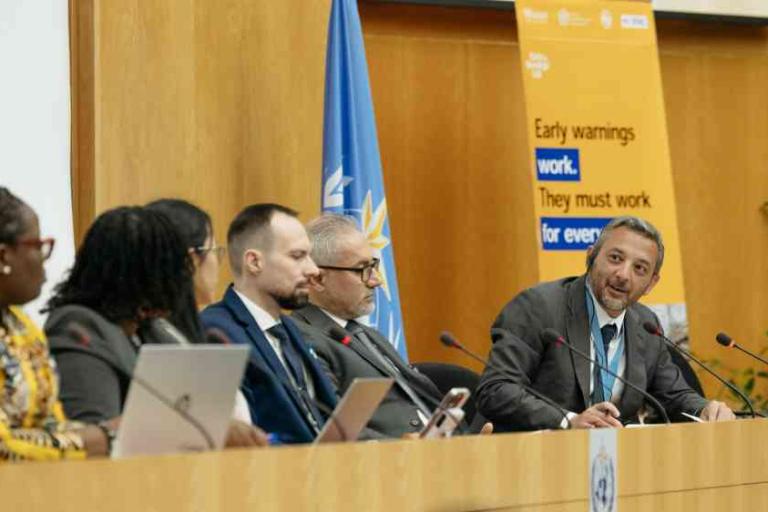 Five people sit at a conference table with microphones, engaging in discussion; a banner behind them reads, "Early warnings work. They must work for everyone.