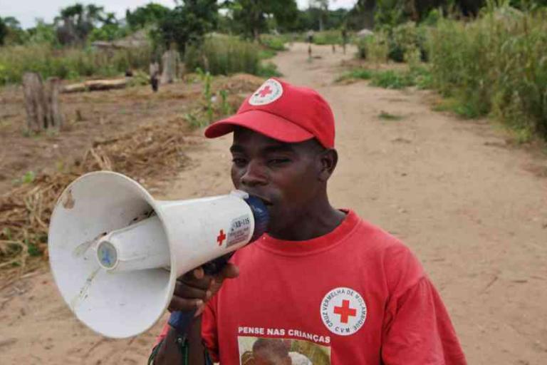 A man wearing a red Red Cross shirt and cap speaks into a megaphone on a dirt path in a rural area.