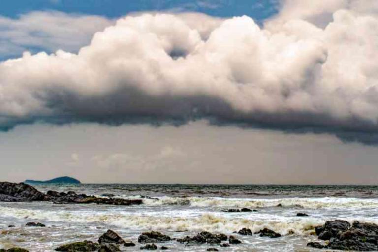 Large, dark storm clouds hang over a rocky shoreline with waves crashing onto the beach and a distant landmass on the horizon.