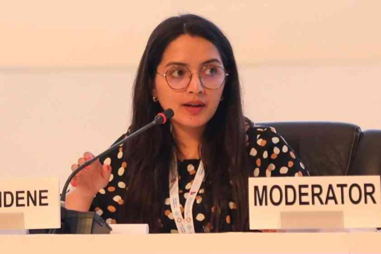 A woman with long dark hair and glasses speaks into a microphone while seated at a panel labeled "MODERATOR." She wears a black top with white spots.