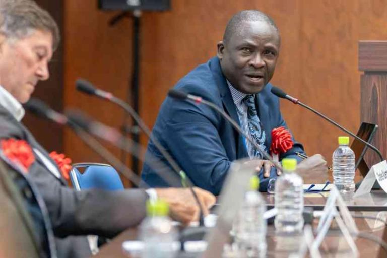 A man in a suit speaks at a conference table with microphones, while another man listens. Bottled water and nameplates are visible on the table.