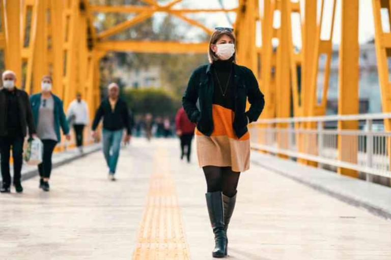 People wearing face masks walk on a yellow-painted pedestrian bridge during the daytime. A woman in front wears a short dress, boots, and a mask. Trees are visible in the background.
