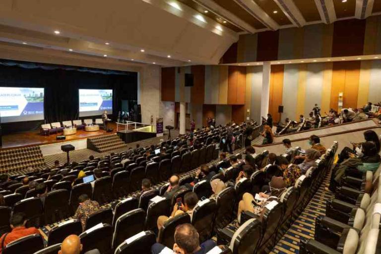 People sit in a large auditorium with tiered seating, facing a stage with screens displaying presentation slides at a conference or seminar.
