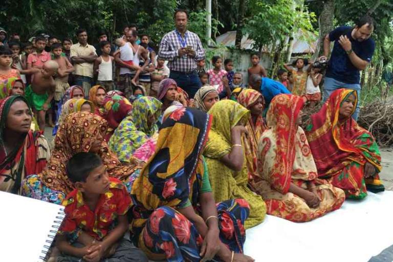 A group of women and children in colorful clothing sit outdoors, while people stand in the background, watching an event or gathering.