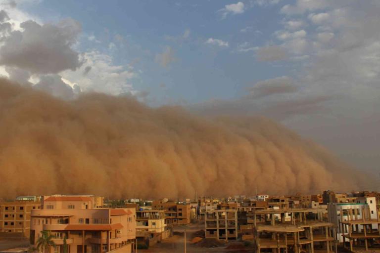 A massive sandstorm approaches a city with residential and commercial buildings under a partly cloudy sky.