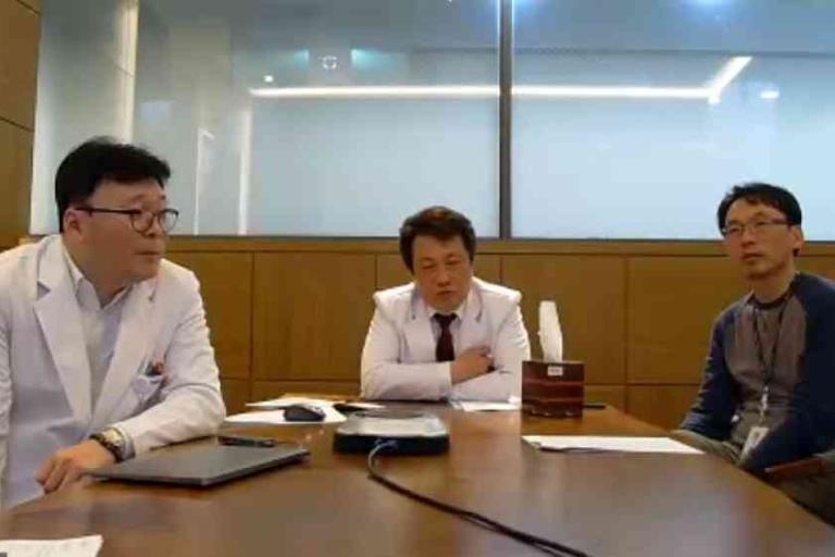 Three men are seated at a conference table in an office; two wear white lab coats, and one wears casual clothes. There are papers and a laptop on the table.
