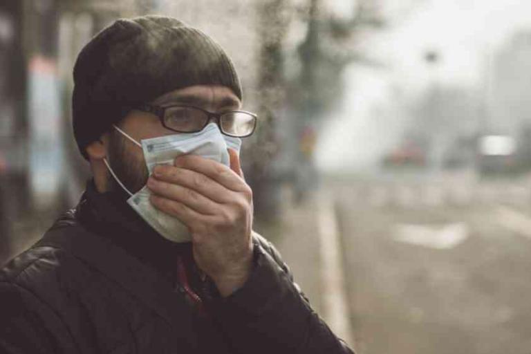 A person wearing glasses and a beanie holds a mask over their face while standing outside on a foggy or polluted street.