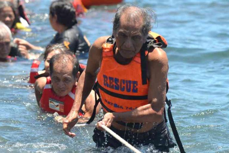 Several people wearing life jackets are pulled through water by a rope during a rescue operation, with one older man leading the group.