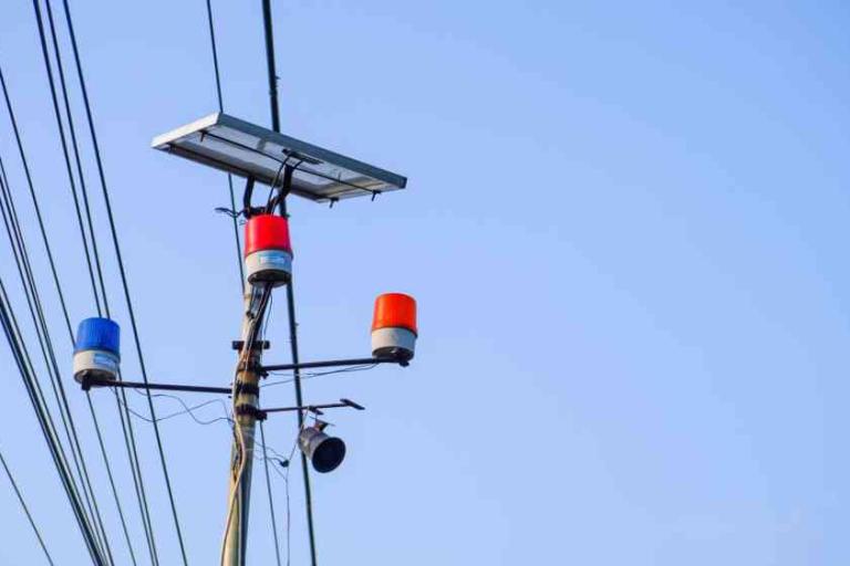 Utility pole with a solar panel, security camera, and red, blue, and orange warning lights against a clear blue sky.