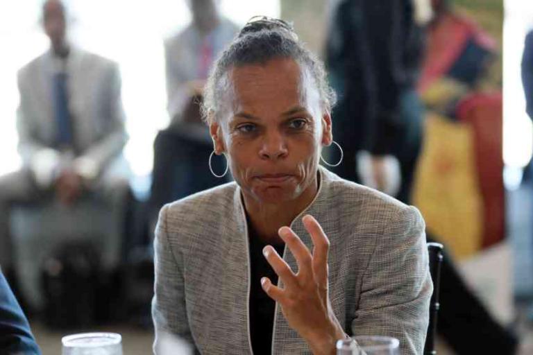 A woman in a gray blazer sits at a table, gesturing with her hand while speaking. Several people are seated in the blurred background.