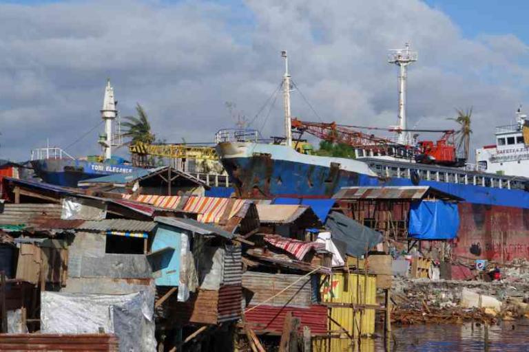 Large cargo ships are docked behind makeshift homes and debris in a coastal area with cloudy skies and scattered palm trees.