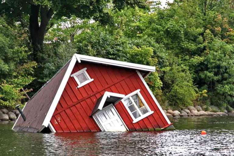 A red wooden house is partially submerged and tilted in a body of water, surrounded by trees and greenery.