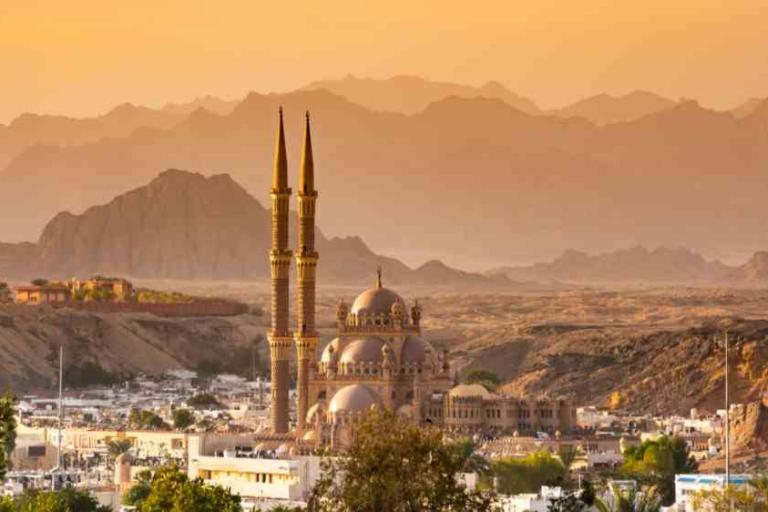 A mosque with two tall minarets stands in a city with desert mountains in the background during sunset.