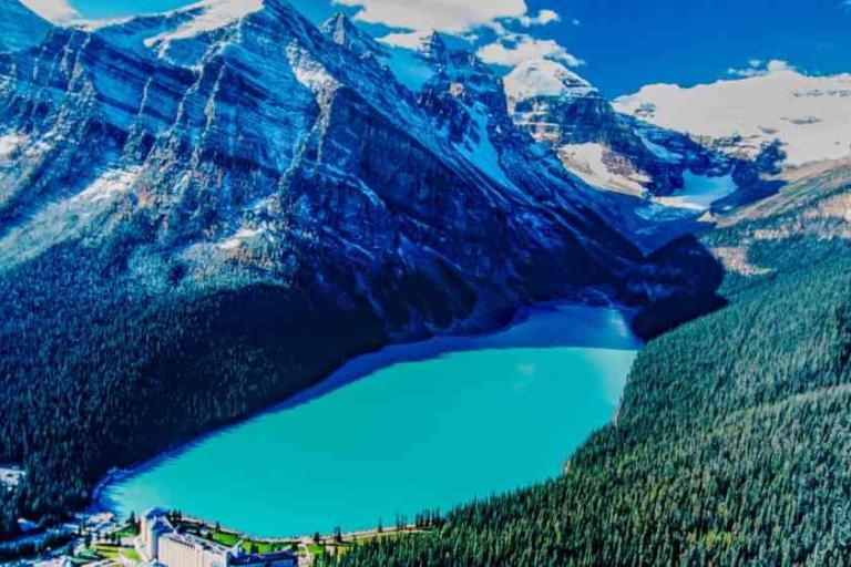 A turquoise lake surrounded by dense forest sits below tall, snow-capped mountains under a blue sky. Buildings are visible near the shoreline.