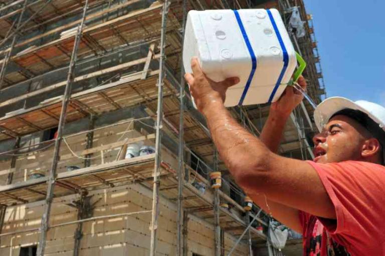 Construction worker in a hard hat lifts a white insulated block in front of a building covered in scaffolding under a blue sky.