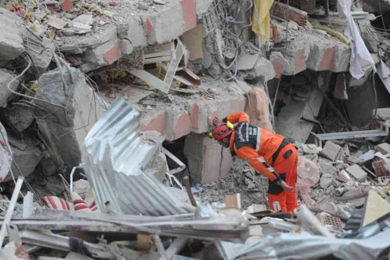 A rescue worker in orange gear searches through debris and collapsed buildings after a disaster, surrounded by rubble and broken structures.