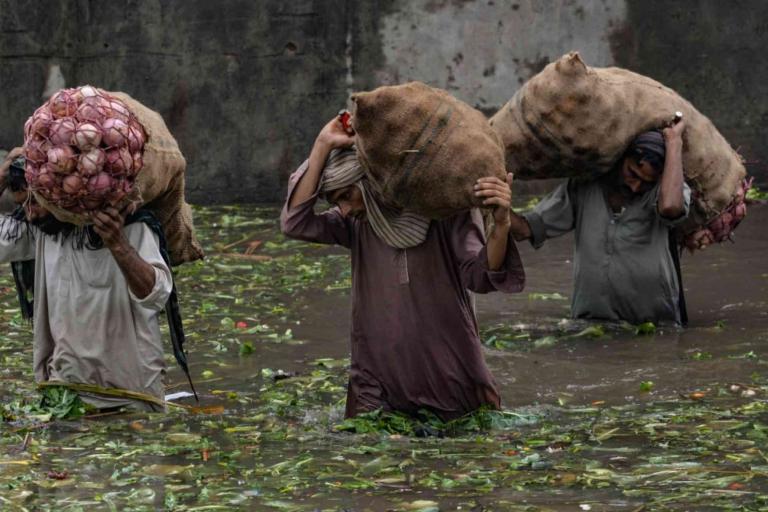 Three people wade through knee-deep floodwater carrying large sacks of vegetables on their shoulders.