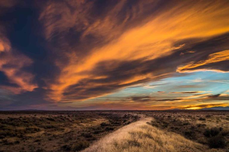 A dirt path winds through dry grass under a dramatic sunset sky with orange and blue clouds stretching across the horizon.