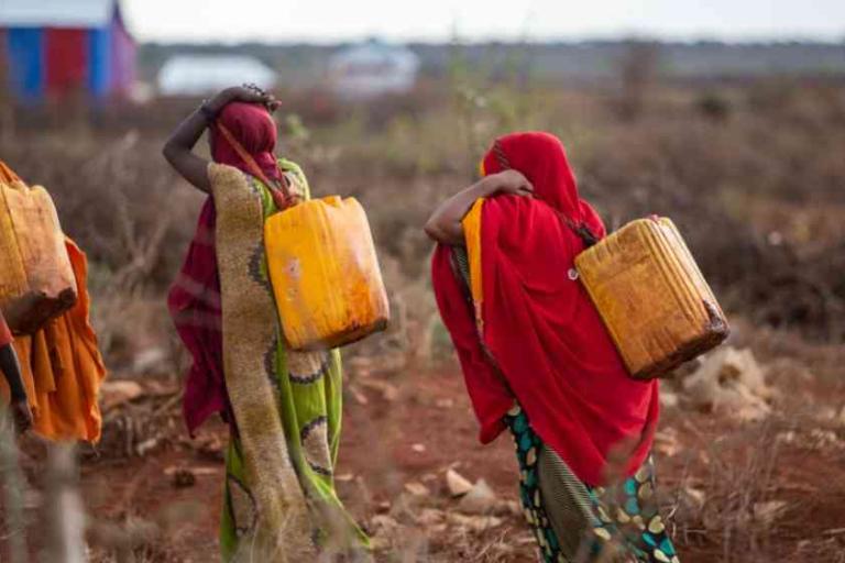 Two women walk through a dry, open area carrying large yellow water containers on their backs, wearing colorful clothing and head coverings.