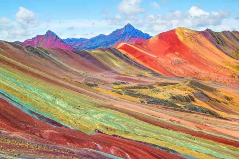 Multicolored striped hills and mountains under a partly cloudy sky, showcasing the natural hues of Rainbow Mountain in Peru.