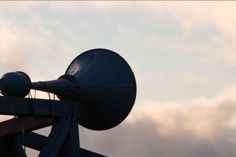 A large outdoor loudspeaker is mounted on a wooden structure against a cloudy sky at dusk.