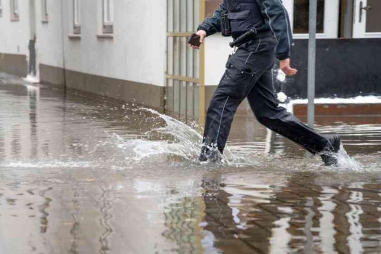 A person in dark clothing walks through a flooded street with water reaching above the ankles in a residential area.