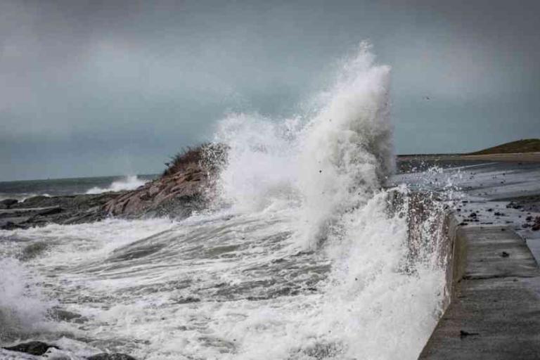 Large ocean waves crash against a rocky shoreline and concrete seawall under a cloudy sky.
