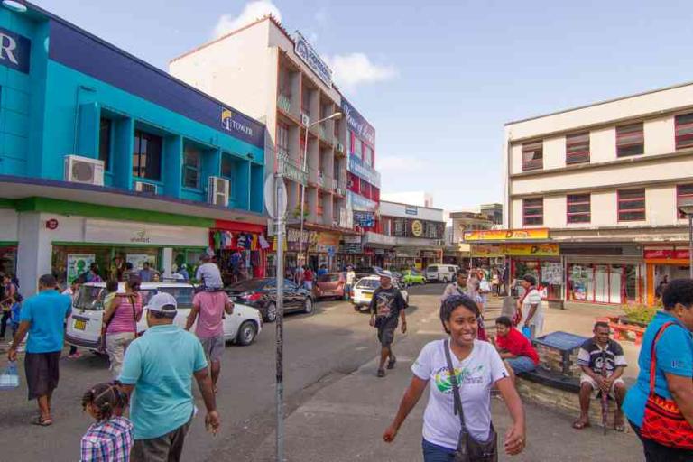 Busy street scene in a town with people walking, colorful buildings, parked cars, and storefronts on a sunny day.
