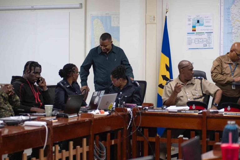 Several uniformed officers and officials sit and stand around a conference table with laptops and documents in a meeting room, with maps and a flag visible on the walls.
