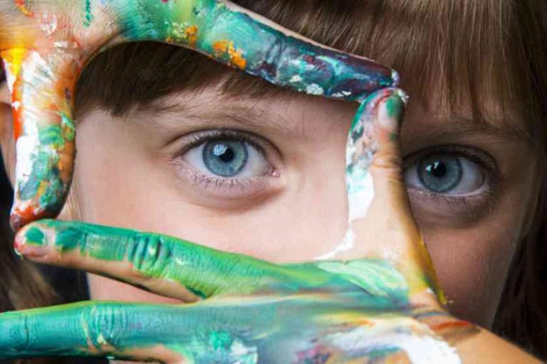 A child with blue eyes frames their face with hands covered in colorful paint, looking directly at the camera.