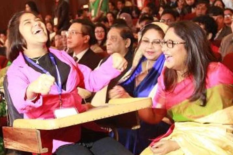 Two women in colorful clothing laugh together in a crowded auditorium, surrounded by people seated and watching an event.