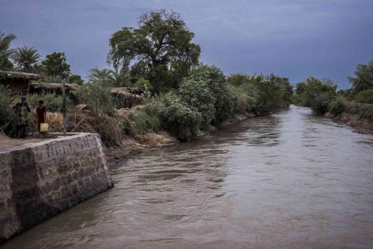 A wide canal with muddy water flows past dense vegetation and simple structures; two people stand on the bank next to yellow jerry cans.