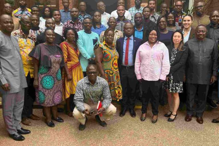A large group of people, both men and women, stand and pose for a group photo on steps outside a brick building.