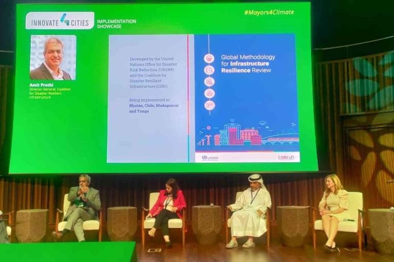 Five panelists sit on stage during a conference presentation, with a green backdrop displaying a project on global infrastructure resilience review.