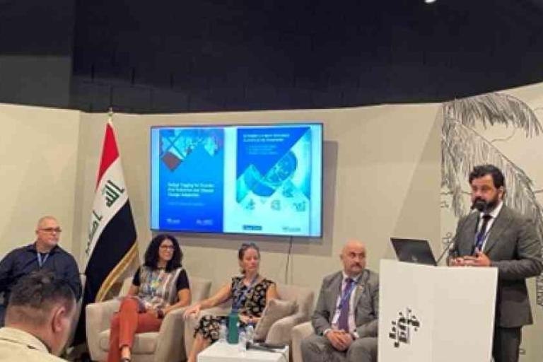 Five people sit on a panel with one man standing at a podium. An Iraqi flag and presentation slides are visible in the background.