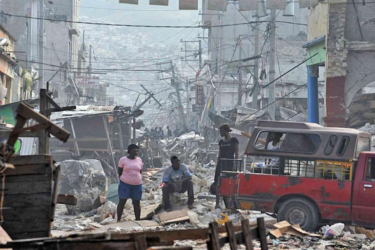 People stand and sit among debris on a street filled with rubble and damaged buildings, with a red vehicle nearby, in the aftermath of a disaster.