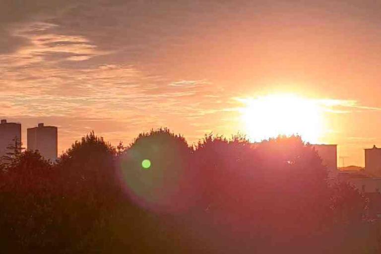 The sun is setting behind trees and buildings, casting a warm orange glow across the sky with a lens flare visible in the foreground.