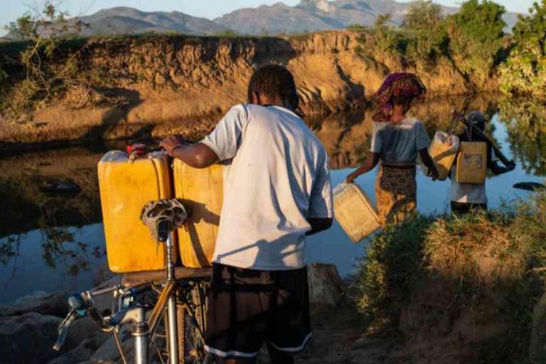 Three people collect water from a river, carrying yellow containers; one person stands beside a bicycle in the foreground. Dry terrain and mountains are visible in the background.