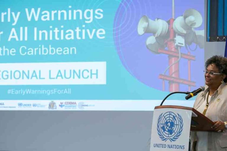 A woman speaks at a podium with the United Nations emblem during the Early Warnings for All Initiative regional launch event for the Caribbean. A presentation slide is displayed in the background.