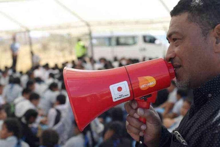 A man speaks into a red megaphone while a seated crowd listens under a white canopy.