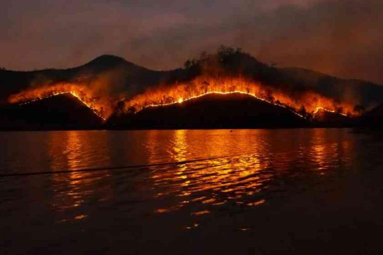A wildfire burns along the ridge of a hill at night, with flames reflected in the water below.