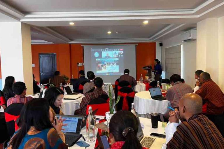 A group of people sit at round tables in a conference room, viewing a presentation projected on a screen at the front of the room.