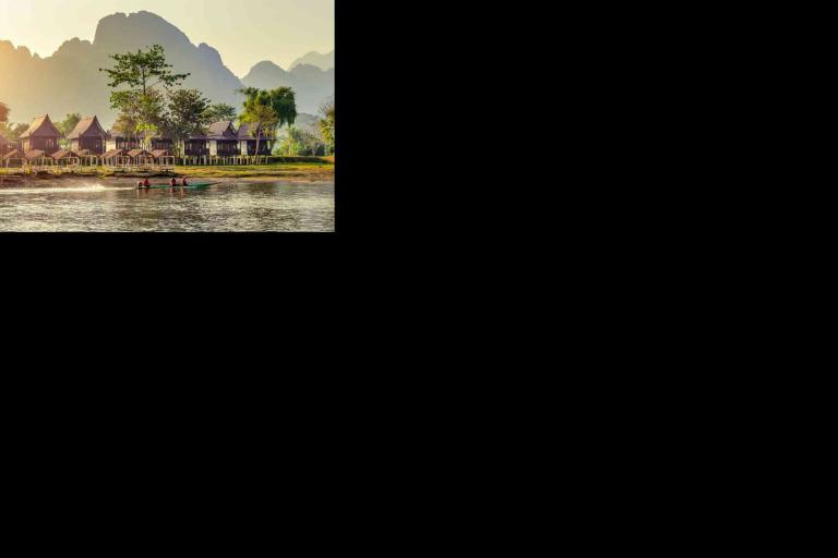 A river with two people in a small boat passes by stilted houses and palm trees, with misty mountains in the background.