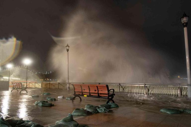 Large waves crash over a waterfront promenade at night, soaking benches and sandbags, with city lights and street lamps visible in the background.