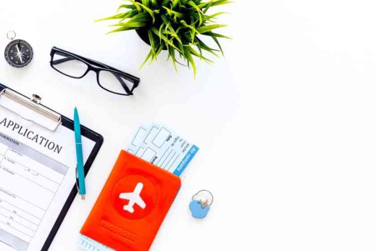 A visa application form on a clipboard is shown with a pen, eyeglasses, a compass, a plant, a passport holder, boarding passes, and a pin on a white surface.