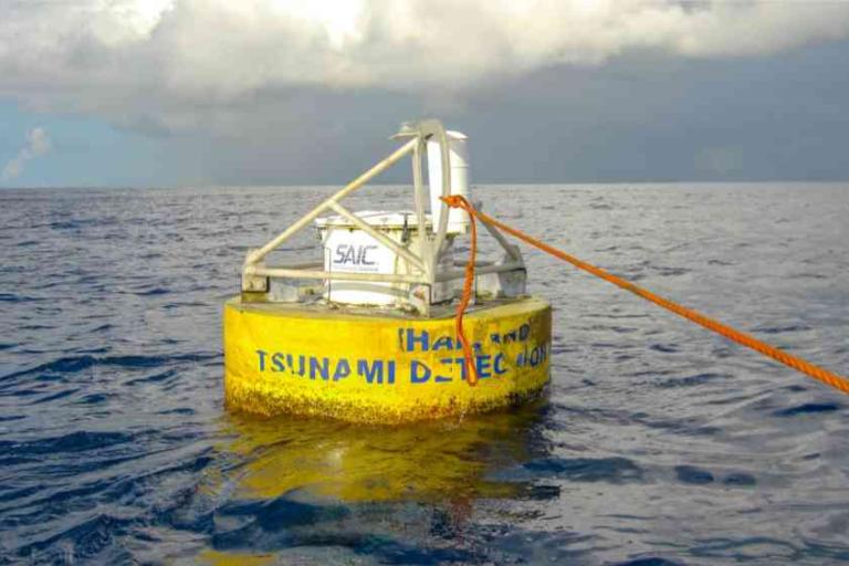 A yellow tsunami detection buoy floats on the ocean, anchored by an orange rope, with cloudy sky in the background.