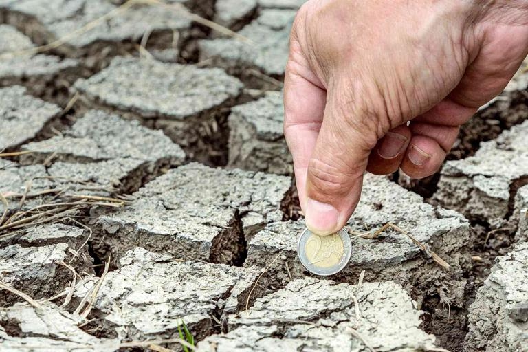 A hand places a one-euro coin into cracked, dry soil, symbolizing investment or loss in drought-affected land.