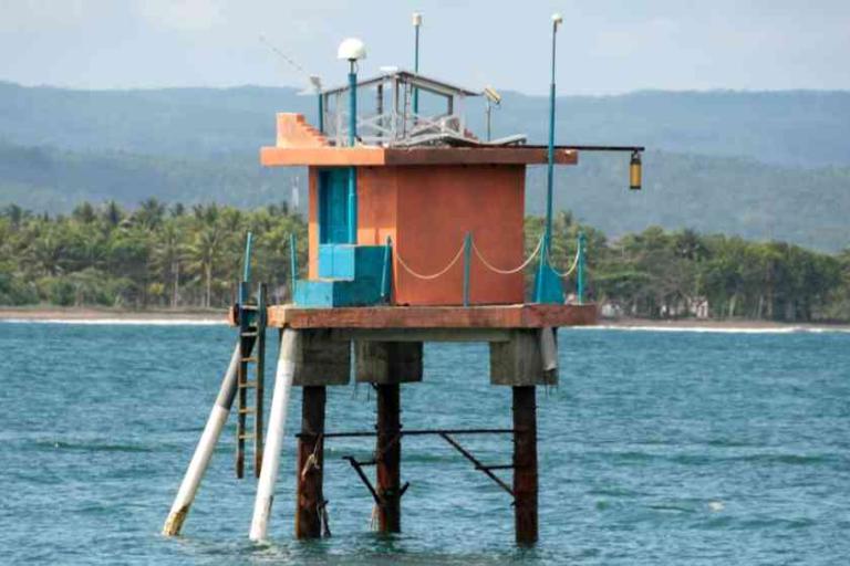 An orange and blue stilted structure stands above the water near a coastline, with a ladder, railings, and equipment on the roof.